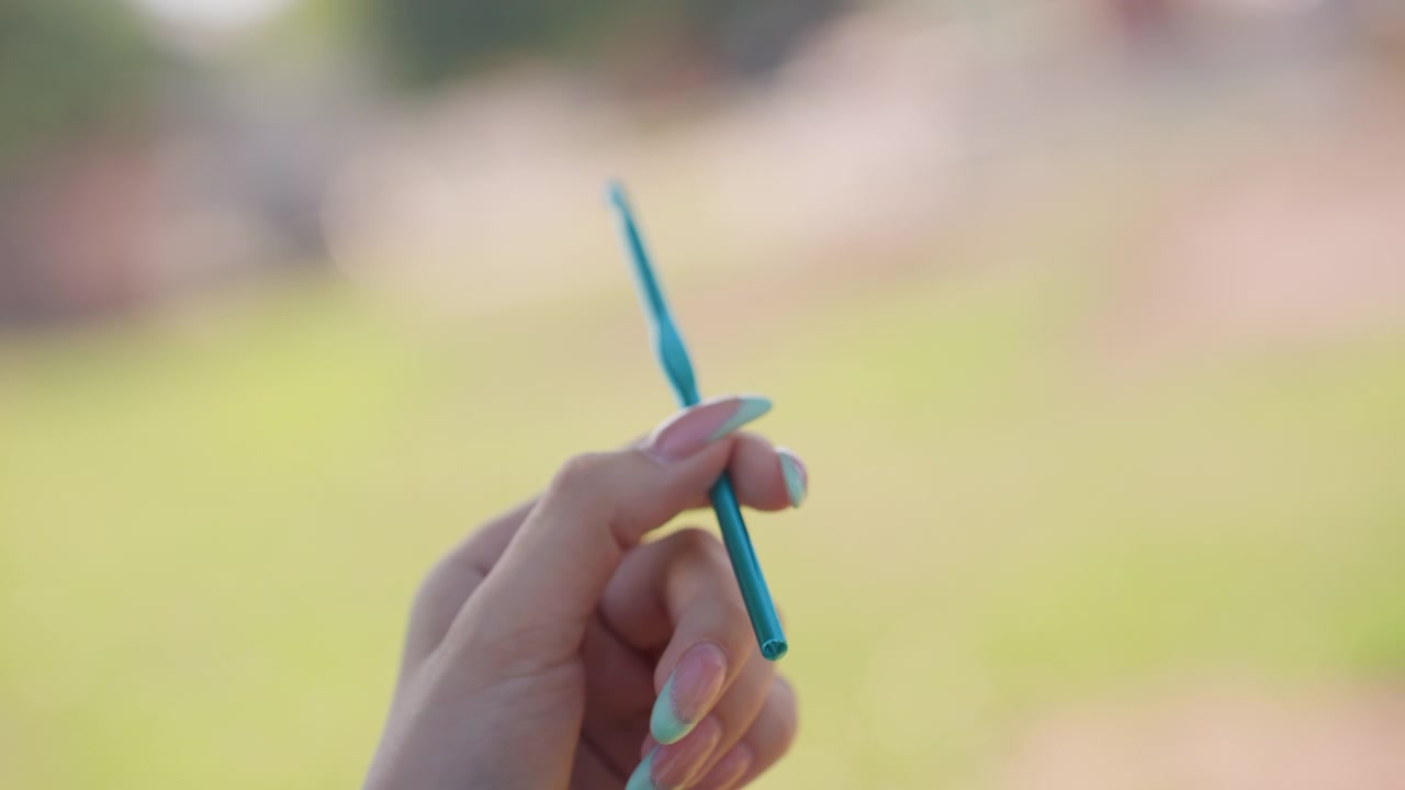 Primo piano di mani con unghie lunghe che tengono e ruotano un uncinetto turchese su uno sfondo verde sfocato. Le dita della donna manipolano lo strumento, mostrandone la forma e la consistenza.
