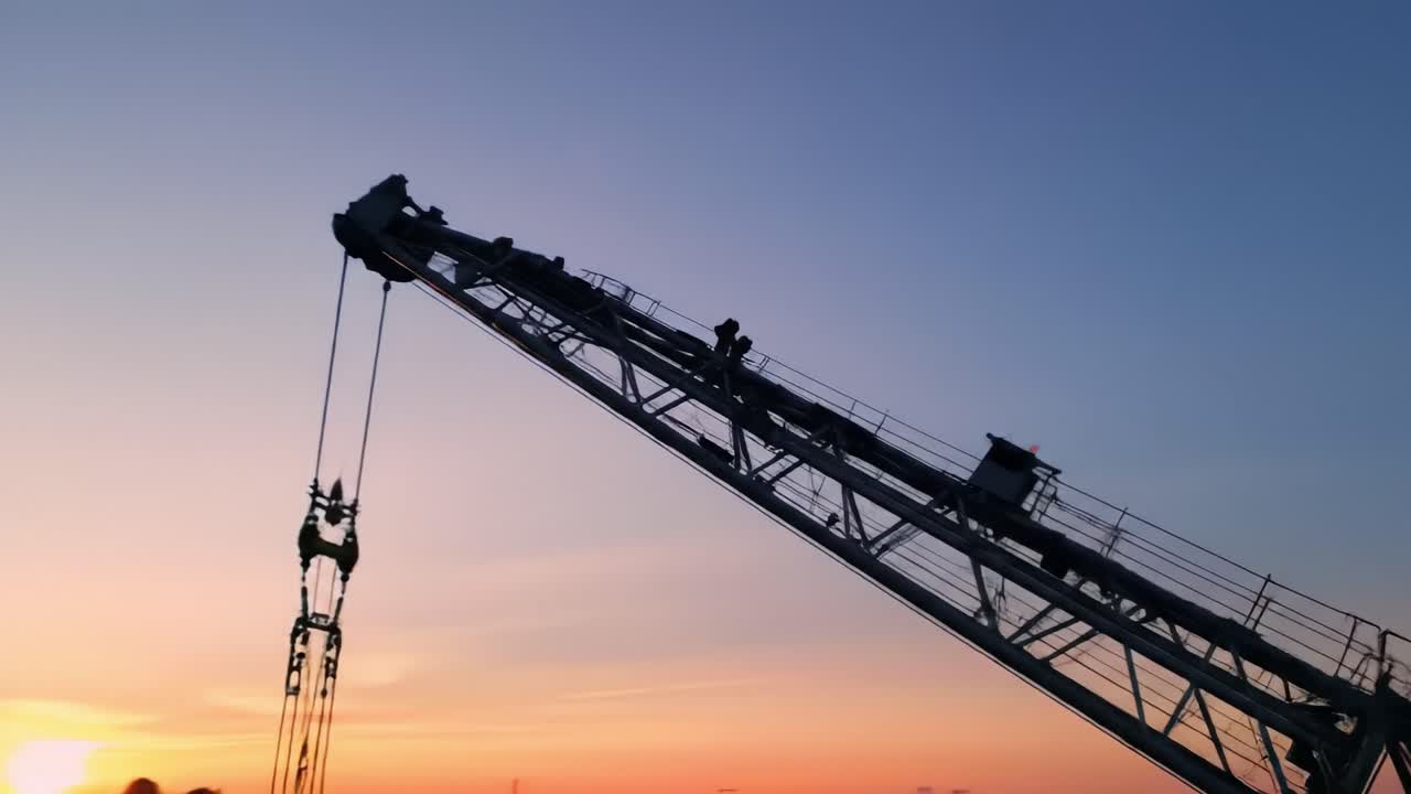 A crane is raised high at dusk, with workers visible on the structure. The skyline reflects beautiful colors as the day transitions to night, highlighting urban development activities.