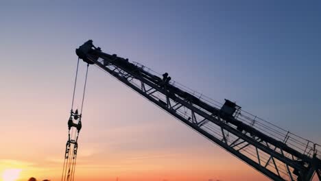 a crane is raised high at dusk, with workers visible on the structure. the skyline reflects beautiful colors as the day transitions to night, highlighting urban development activities.