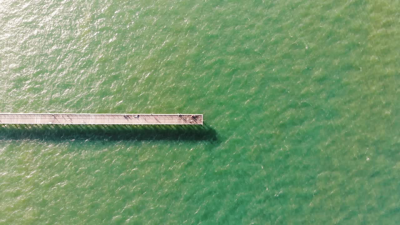 따뜻하고 밝은 태양에서 빛나는 라임 그린과 아쿠아 블루의 색상으로 화려하고 잔물결이 일고 있는 바다에 평화롭게 앉아 있는 긴 목재 부두의 머리 위 사진