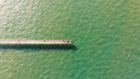 fotografía cenital de un largo muelle de madera que se asienta pacíficamente en un hermoso, tranquilo y ondulante océano con colores verde lima y azul agua que brillan del cálido y brillante sol.
