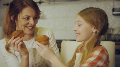 Portrait-shot-of-the-pretty-and-happy-mother-and-daughter-having-fun-while-eating-muffins-at-the-kitchen-table.-Inside
