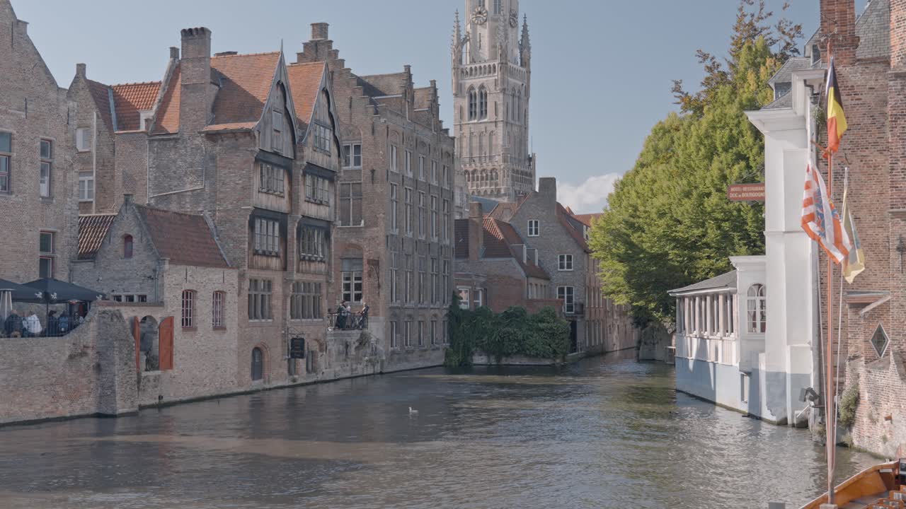A sunny day view along a narrow canal in the historic center of Bruges, Belgium, dominated by the tall, medieval Belfry of Bruges (Belfort) tower towering over the surrounding Flemish gable houses