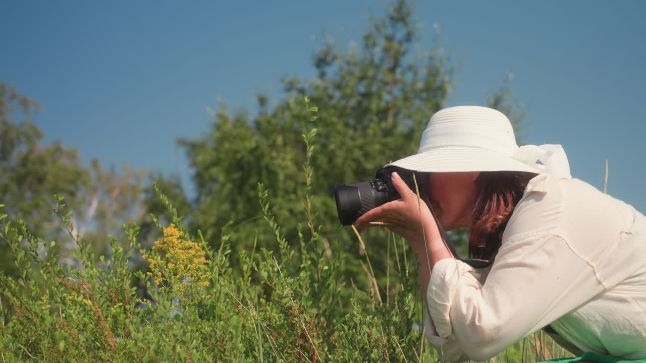 Photographer in wide hat and green dress bends slightly while descending grassy hillside, holding camera and preparing to take closeup flower photo in sunny rural setting with trees
