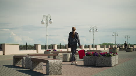 lady in grey clothing walks towards an outdoor bench with flowers nearby, her clothes fluttering in the wind, she drops her shopping bags, sits down, and adjusts her position by crossing her leg