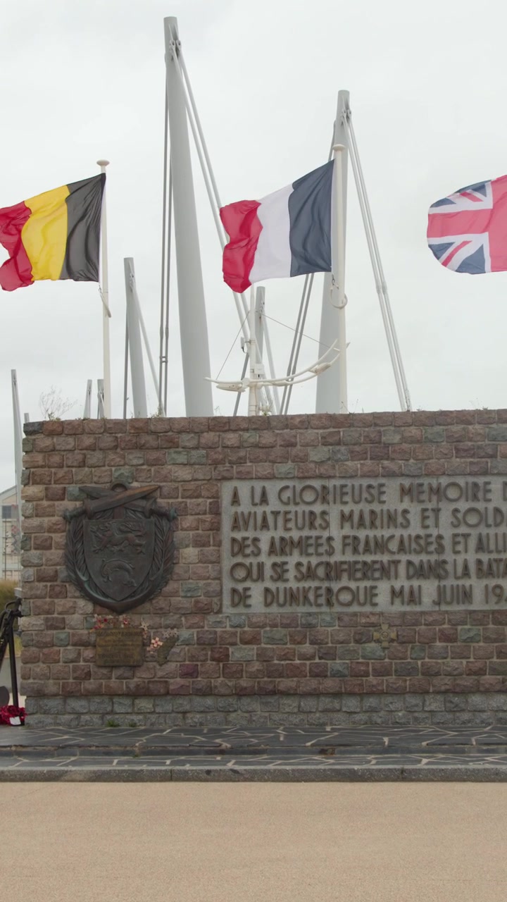 Allied flags flutter above stone WWII memorial, overcast daylight, smooth lateral camera pan, Dunkirk, France