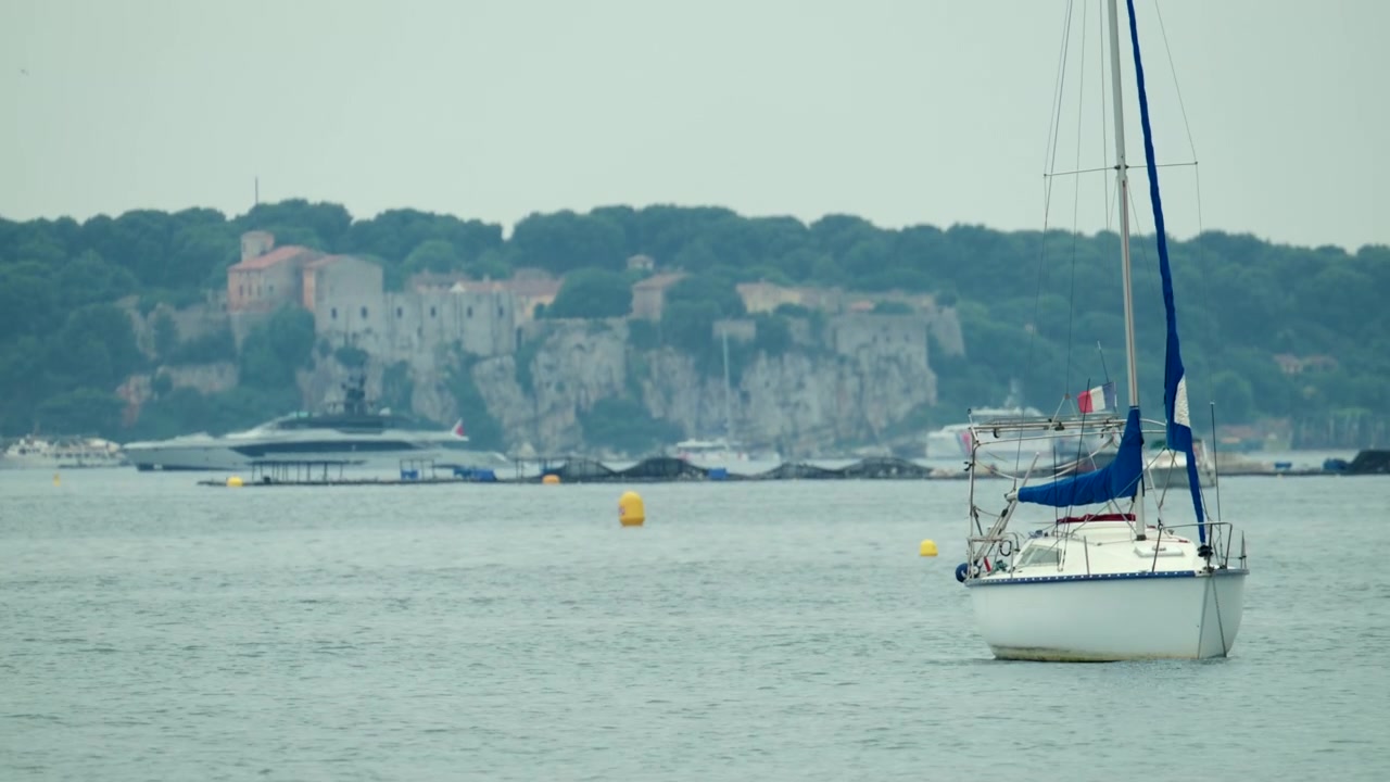 Barca a vela su un mare calmo in una baia, con una lontana città costiera fortificata e un superyacht che suggerisce una destinazione di viaggio di lusso