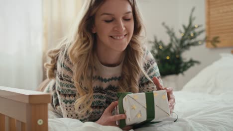 caucasian woman lying down on bed and opening christmas present.