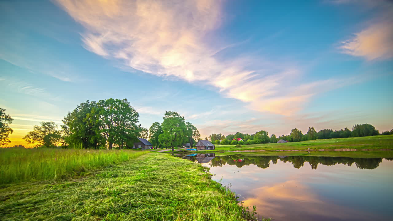 하늘을 가로질러 불고 호수에 있는 시골 별장 위의 물을 반사하는 형형형색색의 일몰 구름 - 시간 경과