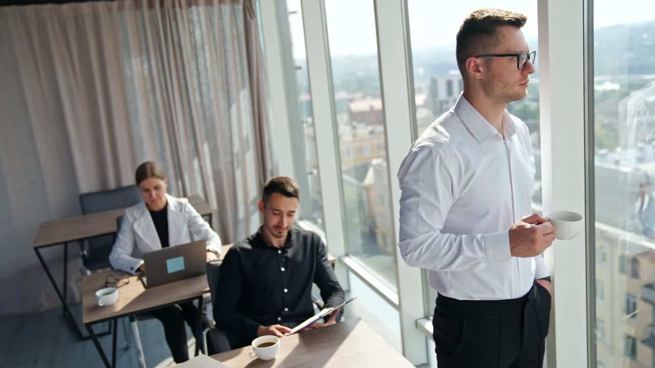 Calm serious businessman in white shirt drinking coffee looking at window. Two employees sitting working at desks.