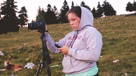 Young-white-woman-in-a-hoodie-preparing-her-camera-on-a-tripod-for-shooting,-with-alpine-meadow-and-pine-trees-in-the-background