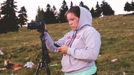 young white woman in a hoodie preparing her camera on a tripod for shooting, with alpine meadow and pine trees in the background