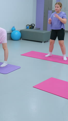 los niños participan en un ejercicio de fitness guiado con un instructor que los conduce en un gimnasio, realizan estiramientos sincronizados en alfombras de yoga coloridas mientras un gran espejo refleja el fondo