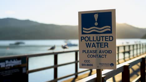 a warning sign highlights water pollution at a scenic pier in akaroa, new zealand, under bright sunlight