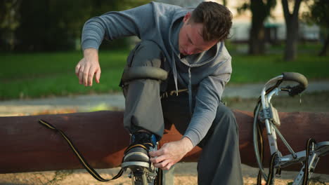 chap sitting on wood log in gray hoodie and dark gray pants, bent over to tighten strap on stilt. another stilt rests beside him on ground. natural outdoor setting with green background