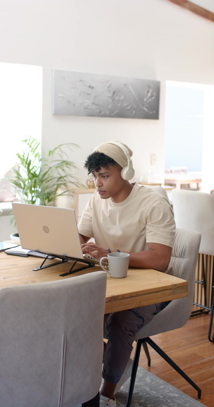 Vertical video: African American man starting work at dining table with headphones typing on laptop