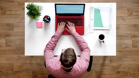 young man in glasses working and typing on laptop, topshot, sitting behind desk with coffee and documents