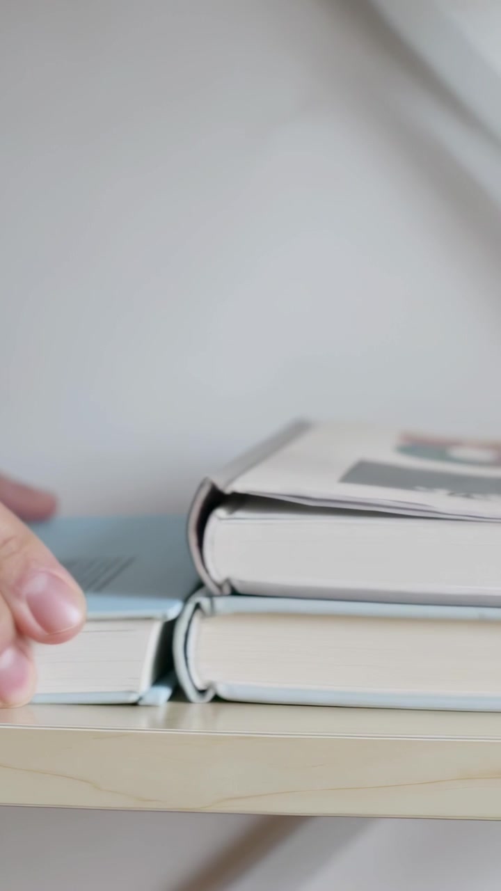 Video verticale: Sollevando un libro con copertina rigida azzurra, mani che fanno scorrere libri su uno scaffale basso di legno allineando i dorsi.