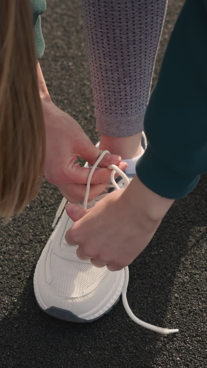 Primo piano di una donna con i capelli al vento che si allaccia le sneakers bianche di tela su una strada asfaltata con segnaletica orizzontale gialla, mettendo in risalto i lacci delle scarpe e l'abbigliamento sportivo