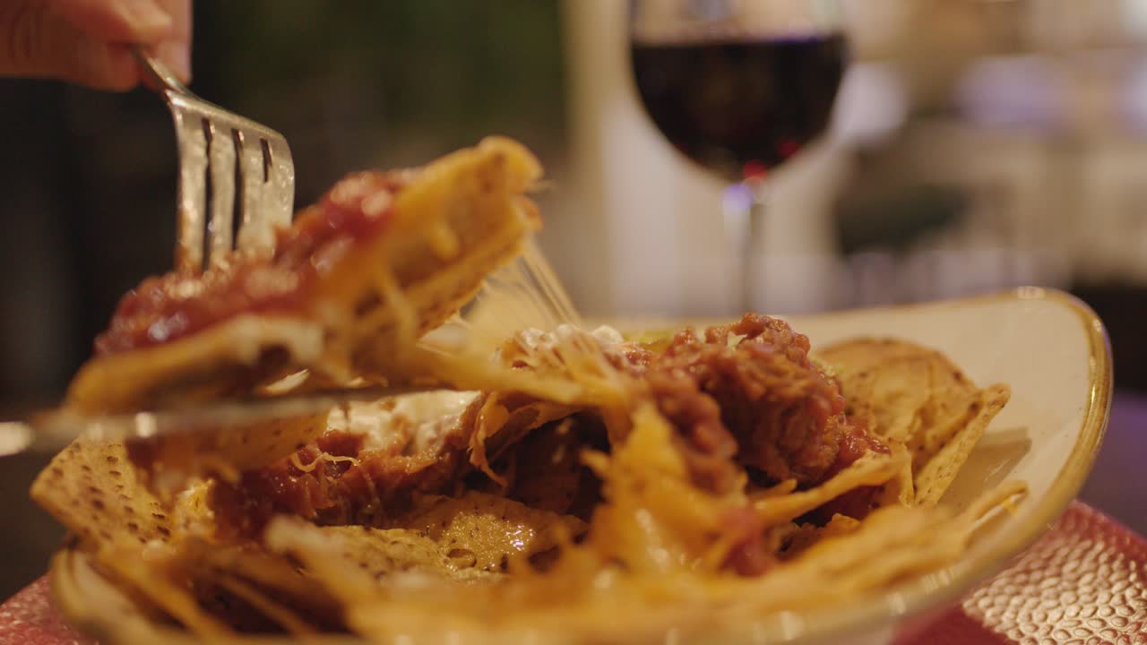 Hand uses fork to pick up loaded nachos, warm lighting, shallow focus, wine glass background