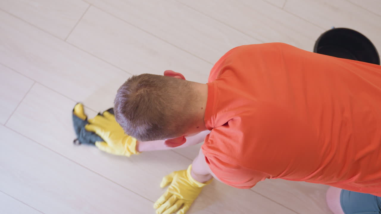 Top down view of man in orange shirt wearing yellow gloves kneeling while scrubbing wooden floor with black cloth, emphasizing hygiene, home care, domestic routine and cleanliness