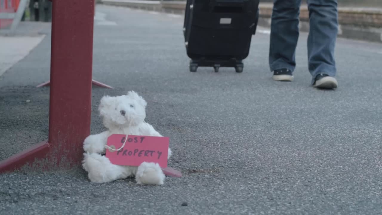 Teddy with lost property tag left on train platform as commuter goes by with suitcase