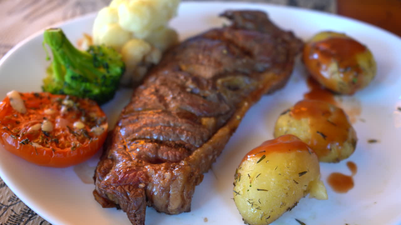 tiro lento estabilizado de bistec a la parrilla en un plato blanco con tomate a la parrilla, brócoli, coliflor y papas al horno
