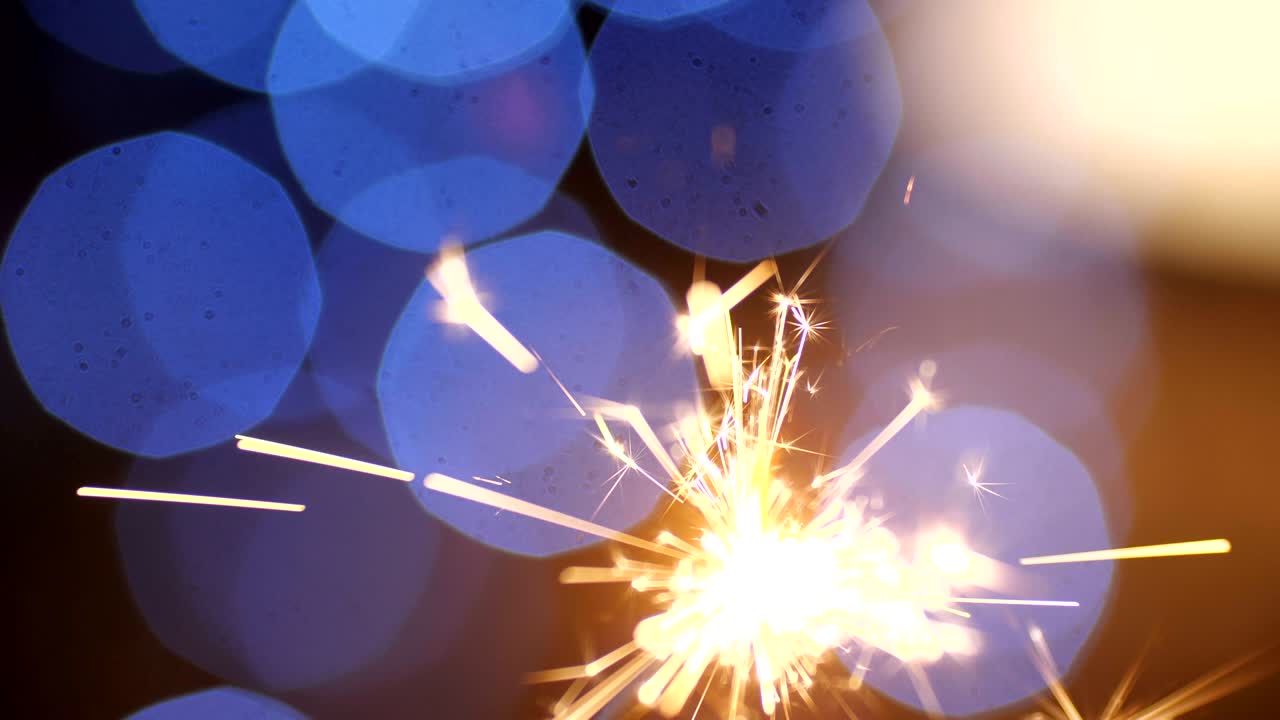 A close-up macro shot of a male hand that setting fire to a New Year sparkler and it is burning, scattering into small sparkles against a background of blue garlands in blur. 4k slow motion