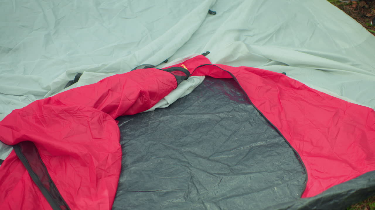 Woman in casual outfit squats on forest floor preparing camp setup while reaching outward, surrounded by dense trees and greenery, as partial leg of passerby becomes visible at frame edge