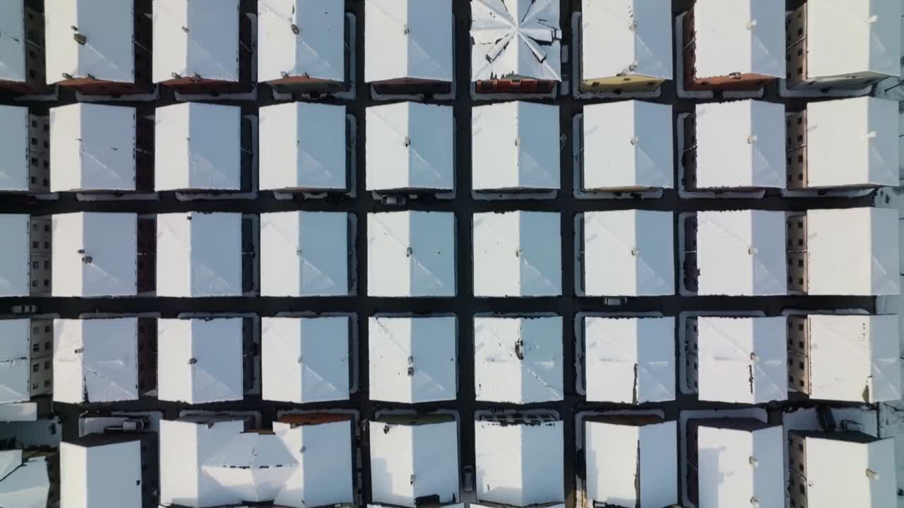 Starting pan, drone moving south over snowy pitched roofs in suburb, revealing parked cars
