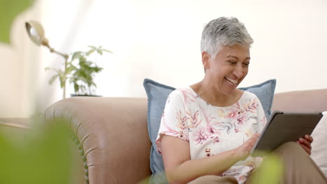 Happy-senior-biracial-woman-sitting-on-couch-laughing-and-using-tablet-at-home,-slow-motion