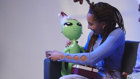 studio shot of young woman wearing reindeer antlers giving christmas present to toy inflatable alien