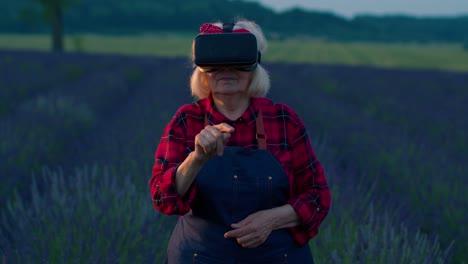 Senior-old-grandmother-farmer-in-virtual-reality-helmet-in-field-of-lavender-flowers-for-management