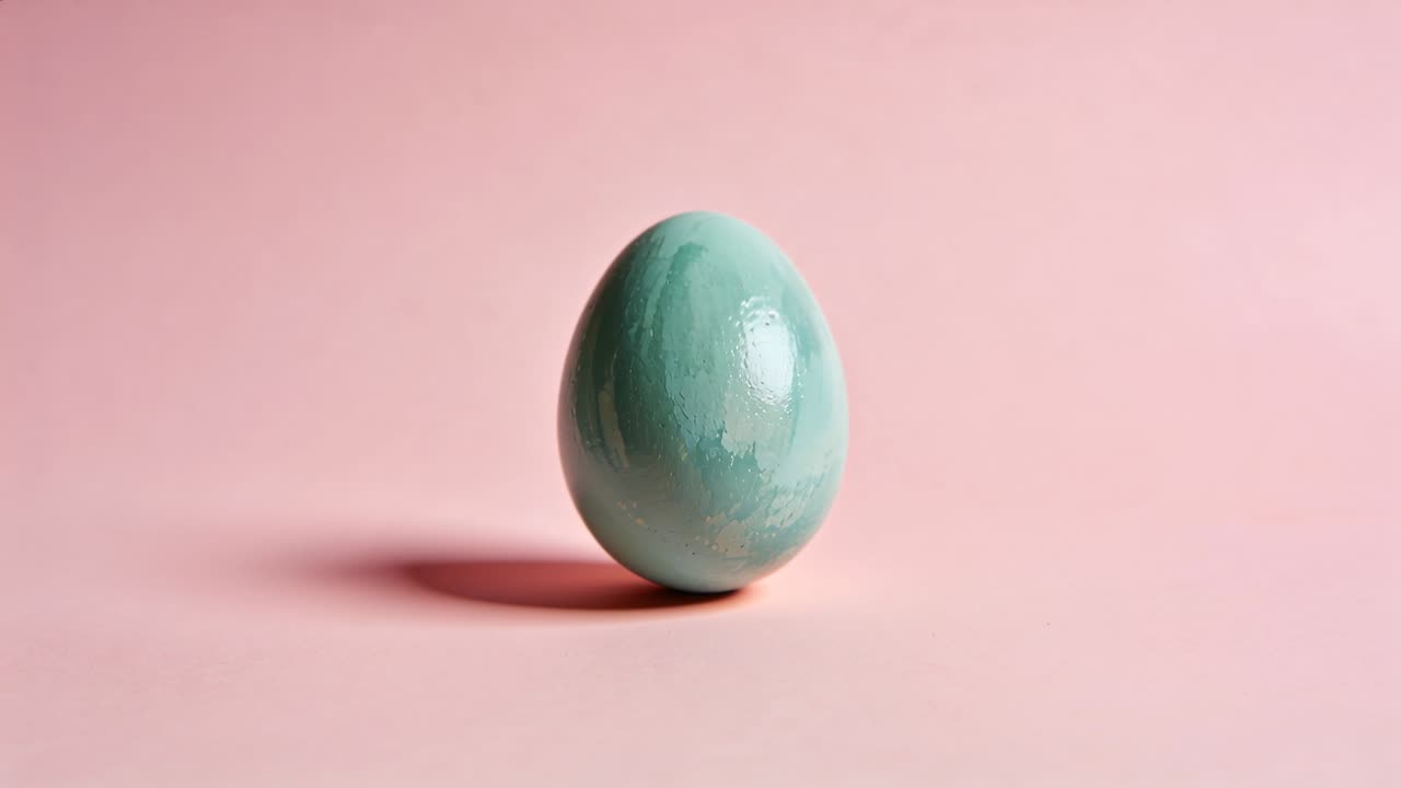 Tilting teal-green painted egg rolling toward right on pastel-pink tabletop, as light shifts