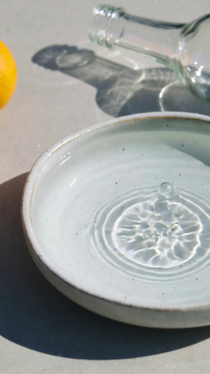 Video verticale: Bottiglia inclinata che versa acqua in un piatto maculato su un tavolo illuminato dal sole, arance.