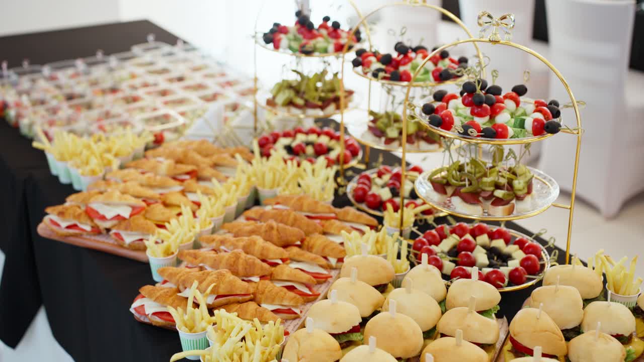 A beautiful and abundant catering buffet table is laden with a delicious assortment of food, including croissant sandwiches, mini burgers, fries, and fresh canapés for a party or event.