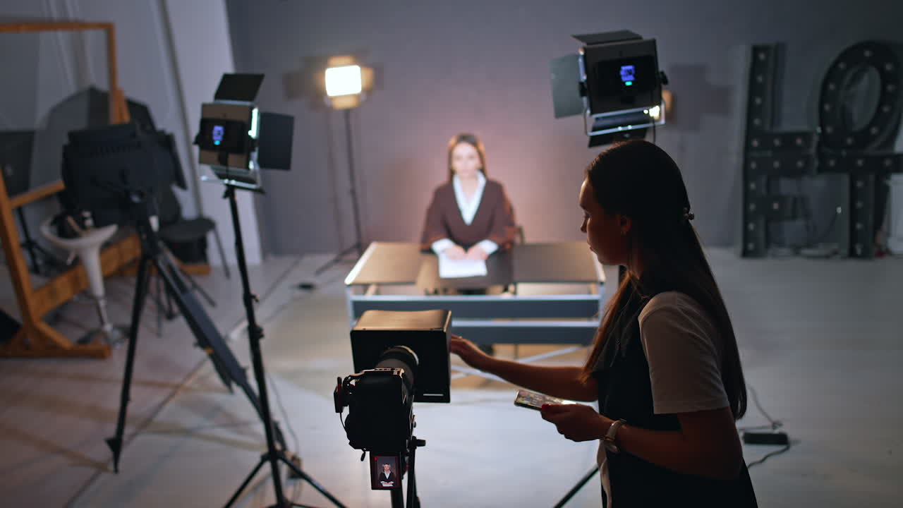 Young long-haired woman with phone in hand stands at camera on tripod touching it. Lady at backdrop sits at desk at blurred backdrop. TV or blog footage in studio.