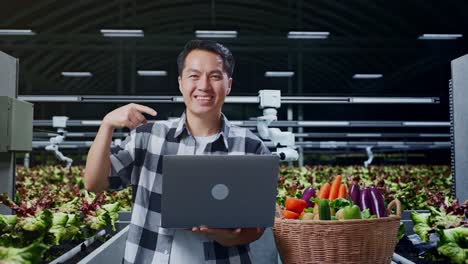 un granjero asiático con una canasta de verduras sonriendo mientras usa y señala una computadora portátil en una granja inteligente