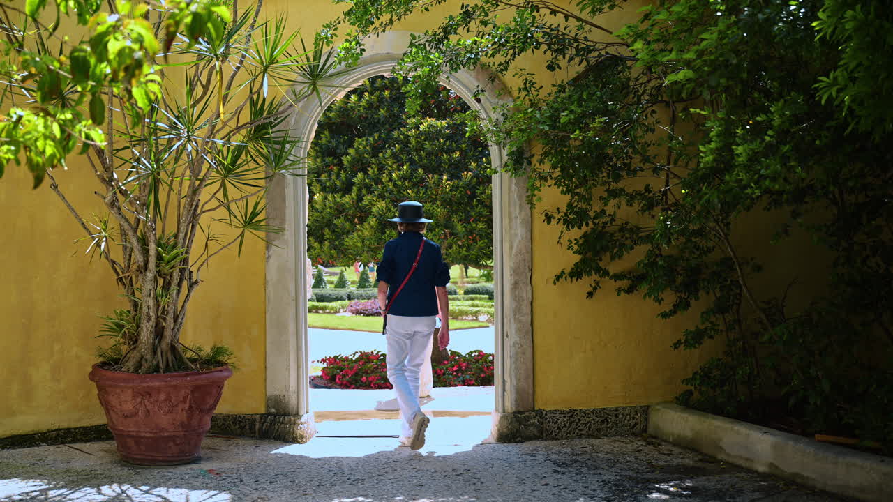 Miami, USA, 6 aprile 2026: Donna che cammina attraverso un portale ad arco in pietra verso un giardino lussureggiante. Vista attraverso un arco in pietra di una donna con un cappello che entra in un giardino tropicale verde brillante a Vizcaya Miami.