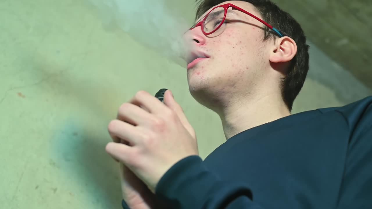 Young pimply man in red glasses vaping and exhales smoke indoor. Side view from the bottom. Close up. Slow motion