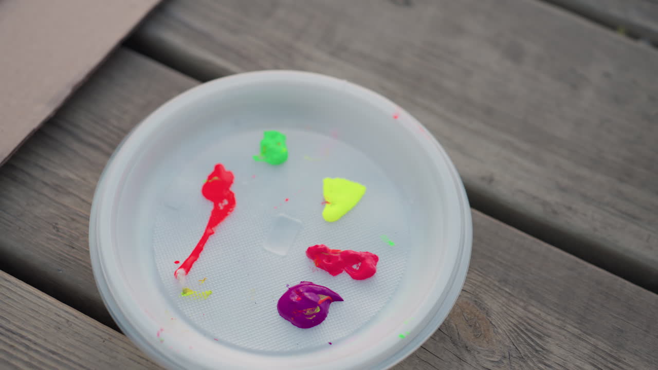 neon red green yellow pink purple acrylic paint dabs in white plastic bowl on weathered wood table outdoors ready for creative art session under soft natural light at riverside park