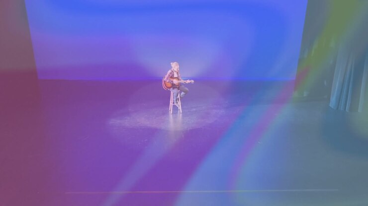 Faretto che si attiva su un musicista che strimpella una chitarra acustica mentre luci arcobaleno si spostano per una promozione musicale.
