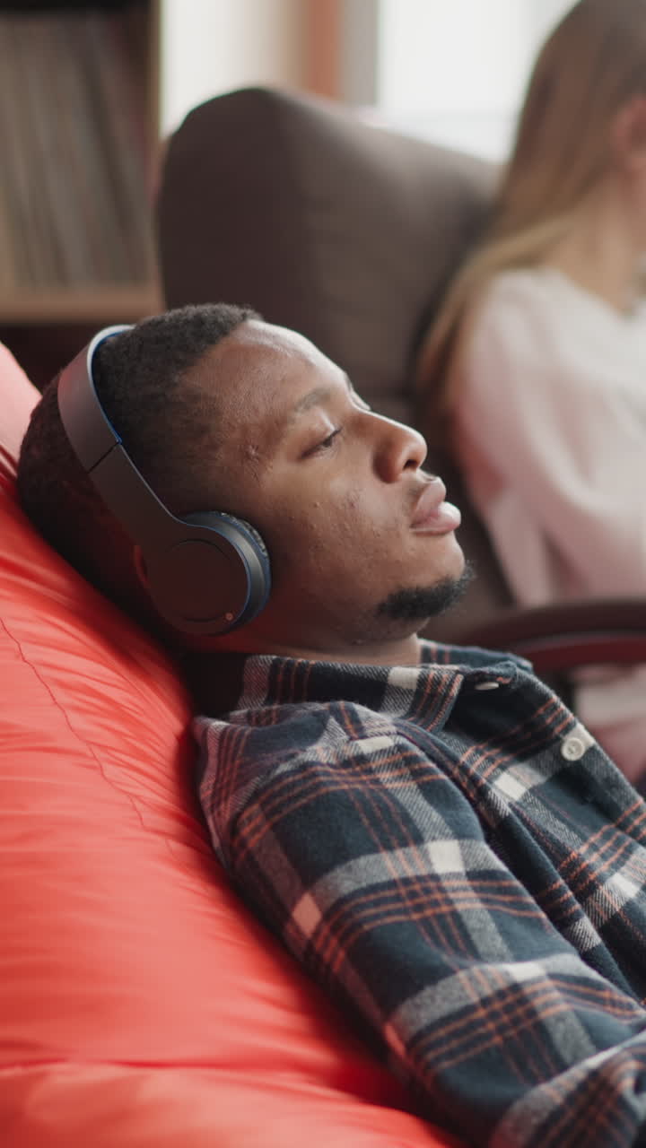 Man listens to audio information via earphones in library. Black guy with headphones sits on soft bean bag resting in cozy bookstore. Nerd leisure