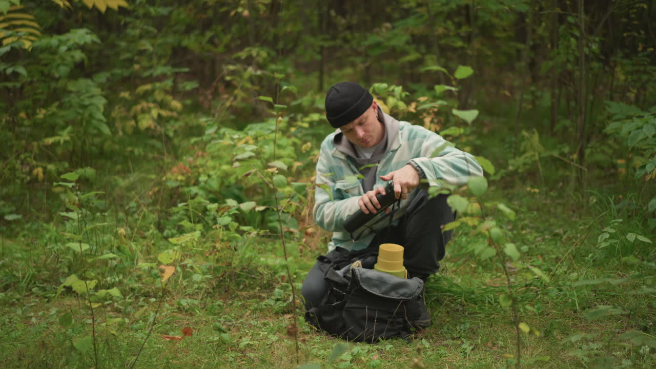 Man die met een mes een houten stok snijdt nabij een rugzak op een open plek in het bos, schaaft krullen af terwijl hij de balans test, maakt een wandelstok of aanmaakhoutgereedschap, meditatieve ambachtelijke buitenscène vol focus