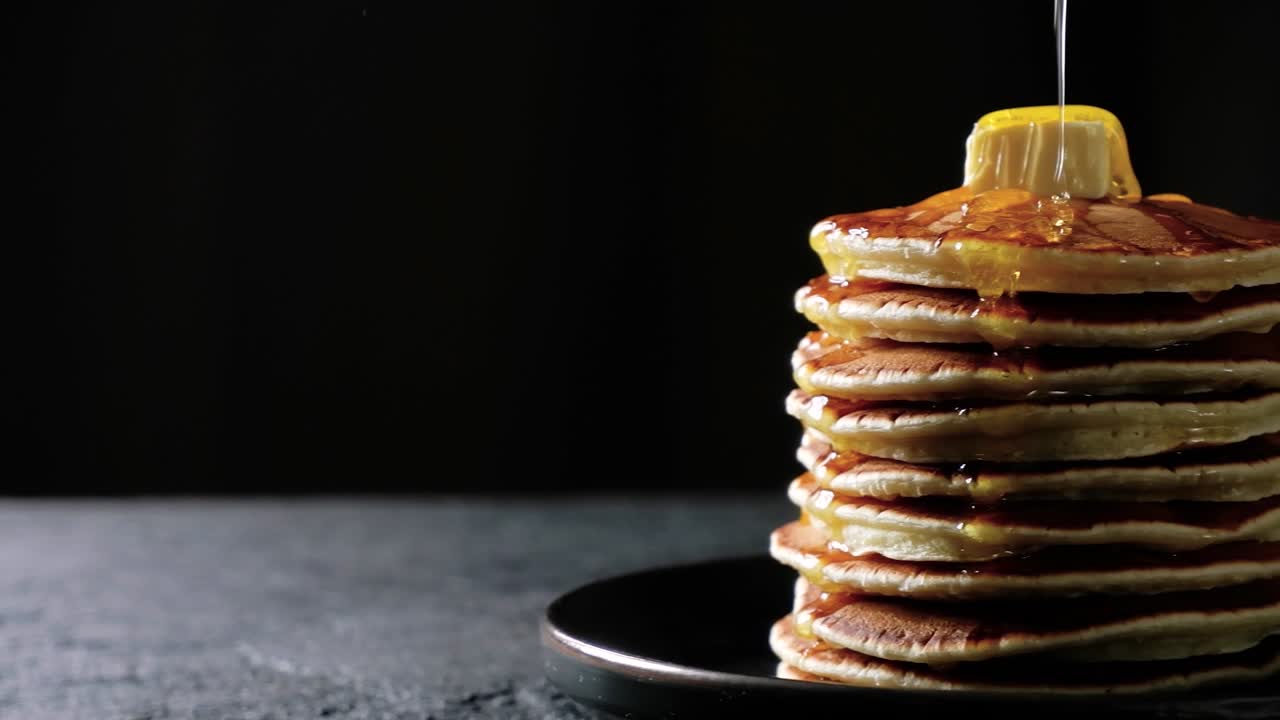 la miel o el jarabe de arce se vierte sobre una pila de panqueques con mantequilla moviéndose sobre un fondo oscuro. imágenes de alta calidad 4k