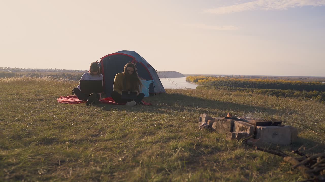 태블릿과 노트북을 가진 소녀들이 강에서 캠프에서 텐트에 앉아 있습니다.