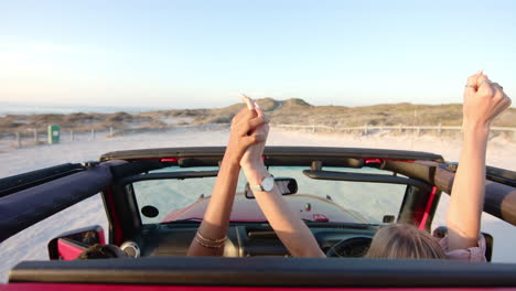 dos personas levantan sus manos alegremente en un convertible rojo cerca de la playa en un viaje por carretera