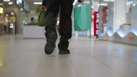 man legs walking through shopping mall polished tile floor wearing black pants and casual shoes holding green shopping bag with blurred storefronts bench and seated shopper in background