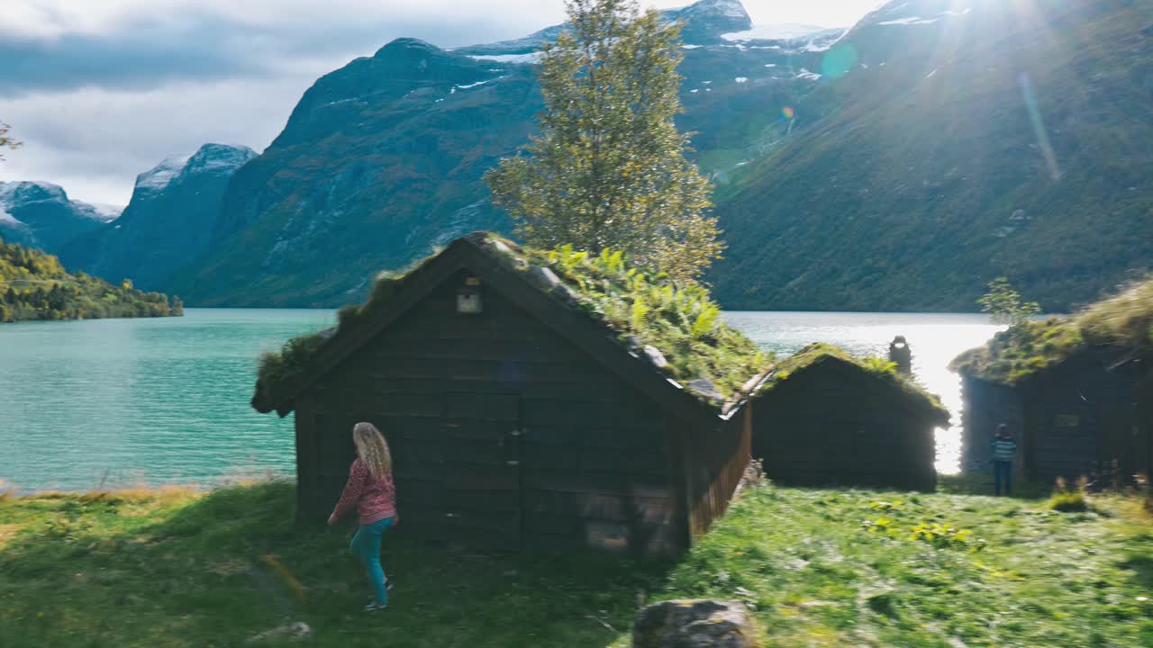 Camera slightly panning and following a girl walking through an abandoned old village, watching over a scenic lake and mountain view in Loen, Norway