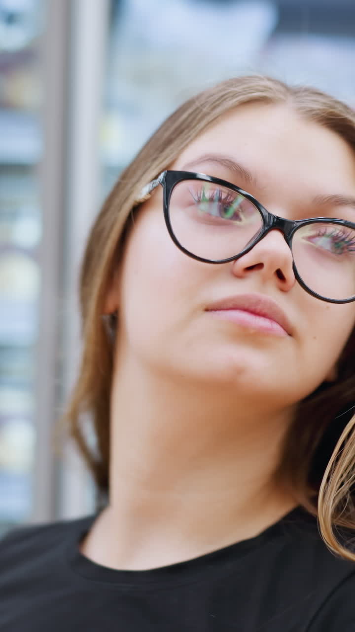 vista lateral de primer plano de una mujer con camiseta negra y gafas caminando en el centro comercial, mirando a su derecha a las mercancías expuestas detrás del vidrio, la iluminación brillante y la expresión enfocada mejoran su experiencia de compra